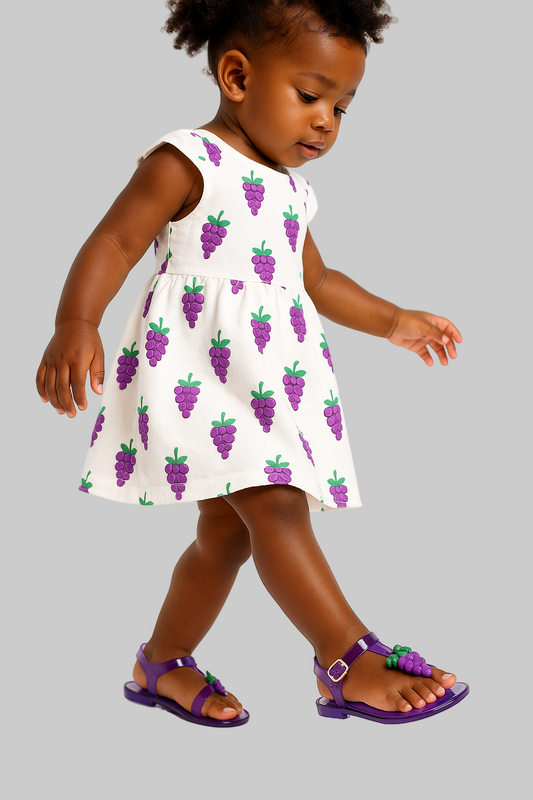 Child wearing a white dress with purple grape pattern and matching sandals on a white background