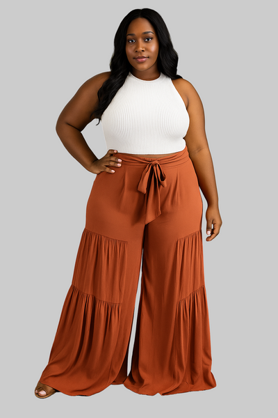 Woman wearing a white sleeveless top and rust-colored tiered pants on a gray background