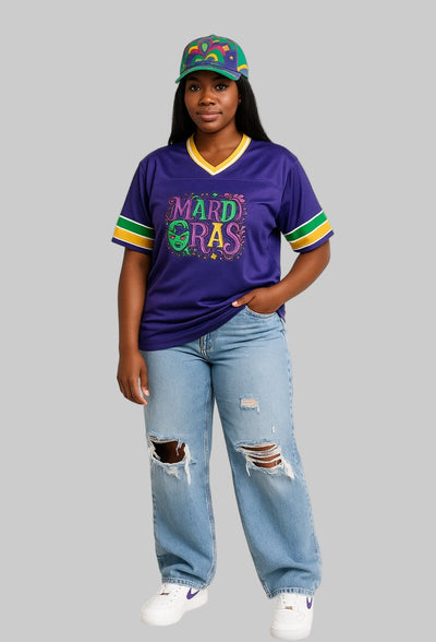 Person wearing a purple 'MARDI GRAS' jersey, light blue jeans, and a colorful cap on a white background