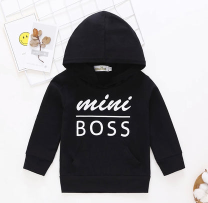Black or white sweatshirt with the text 'MINI BOSS' printed on the front hoodie with a hood, displayed on a flat surface with decorative items around it.