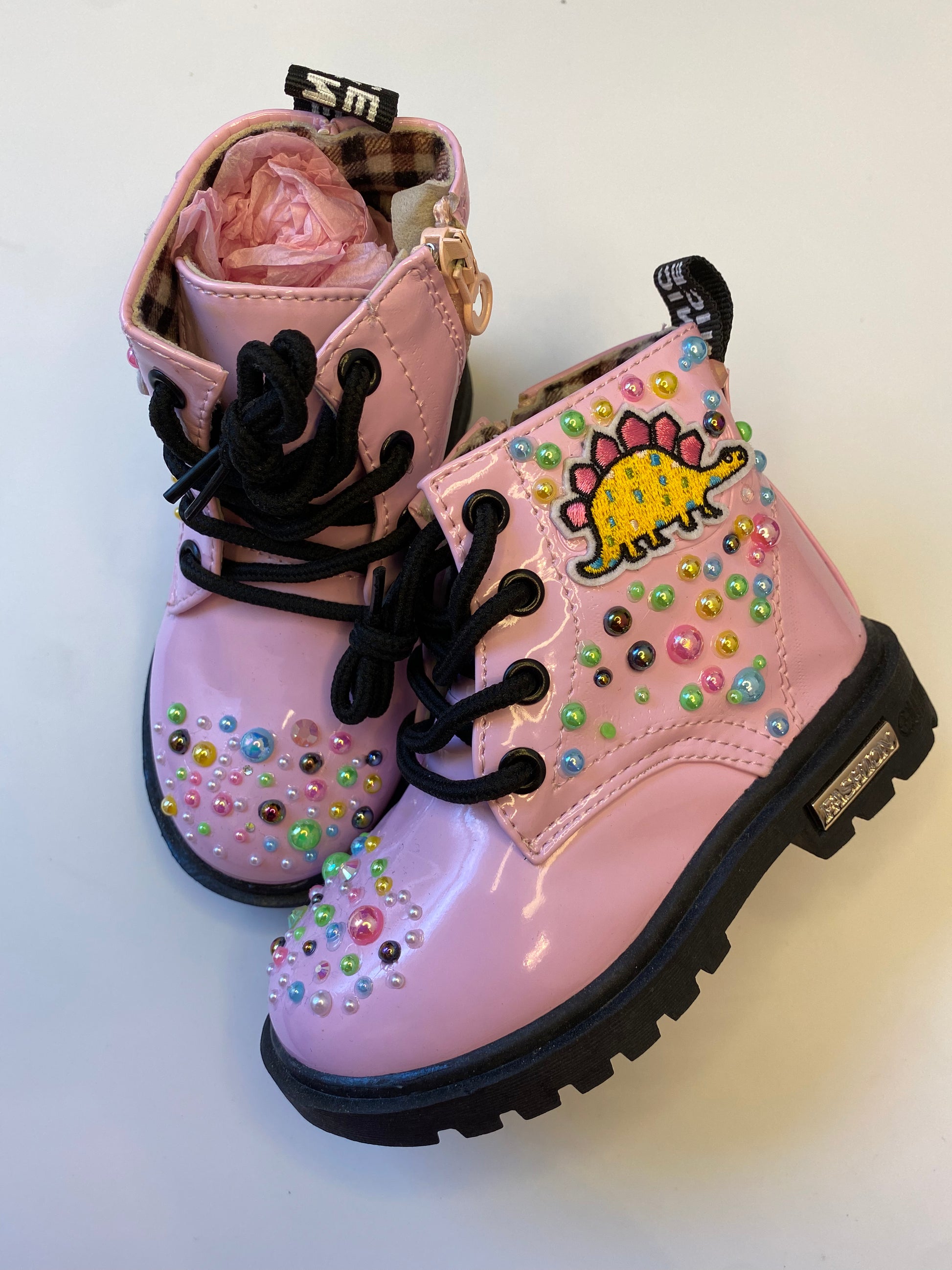 A pair of glossy pink toddler boots with colorful rhinestone embellishments and a playful yellow dinosaur patch on the top, with black laces and soles.