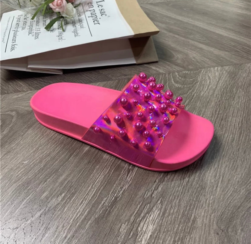 A pink open-toe summer slipper with a translucent strap and pearl decorations, placed on a wooden floor next to a book and a flower.