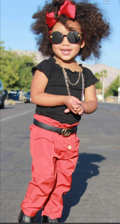 A toddler girl wearing a red and black two-piece outfit with off-shoulder sleeves, flare details, and a chain belt.